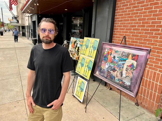 Jerin Sage, project organizer, stands with sunglasses and hands in pockets in front of art displays on sidewalk