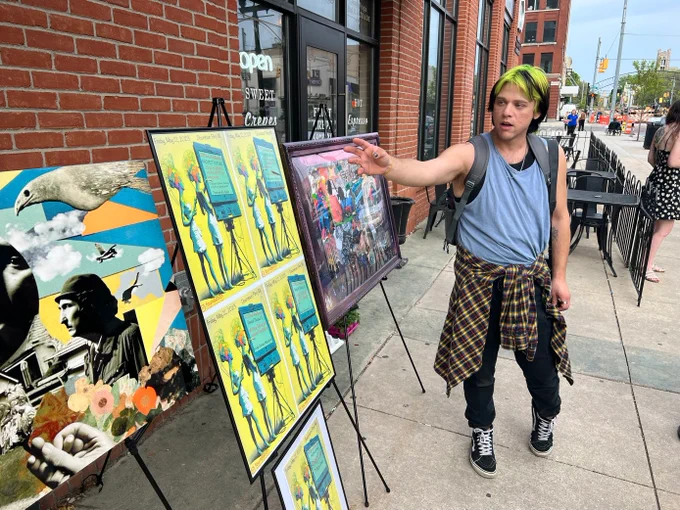Artist (Robby Lee) wearing a grey tank top, a flannel shirt tied around their waist, dark jeans points at art posters in a collage style. 
