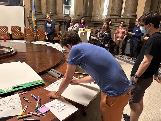 A man in a t-shirt leans over a giant pad of paper to write ideas as other mill about.