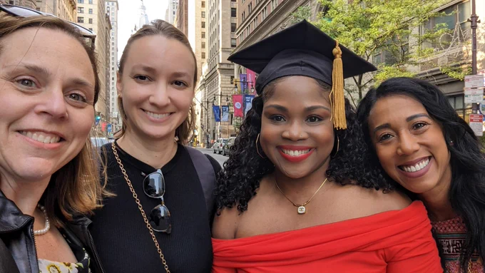 Four women standing on a sunny street in Philadelphia. One is wearing a graduation hat, celebrating her achievement. They appear to be joyful. The vibrant cityscape serves as a backdrop.
