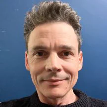 A smiling male looks straight at the camera against a blue background and wearing a black coloured top just visible.
