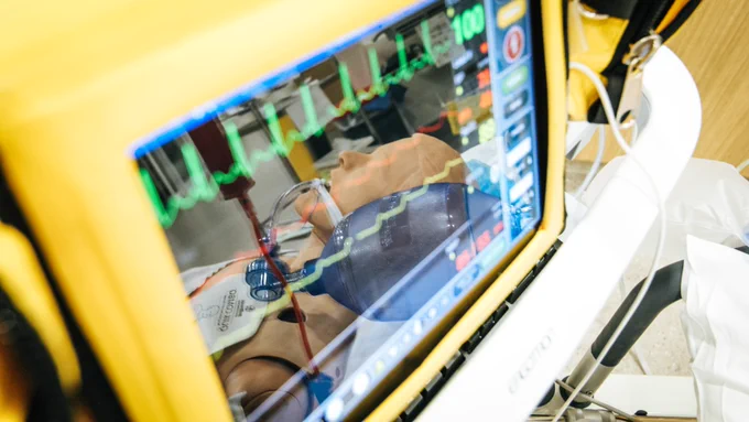 Image of vitals screen, with the reflection of a patient intubated and lying on a hospital bed - only the patient is a mannequin, beige-white in colour and gendered male.