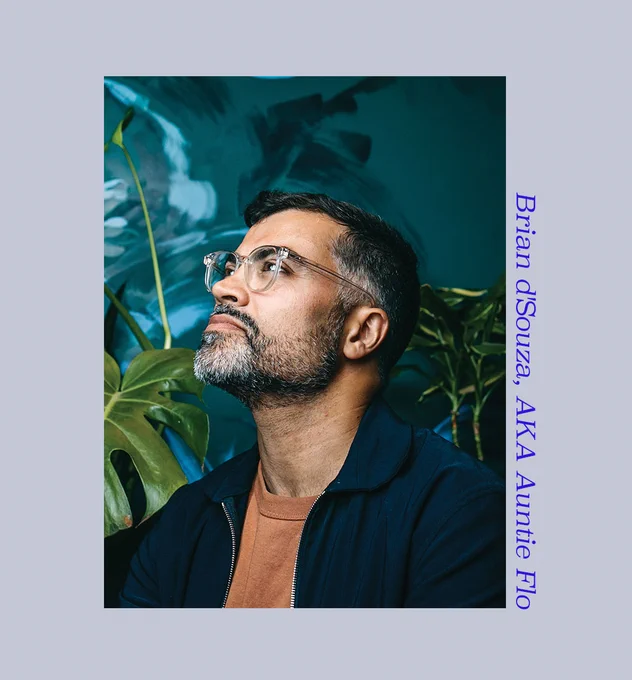 Photo of Auntie Flo wearing a dark blue coat and orange t-shirt. He's on front of a blue background with some green plants behind him