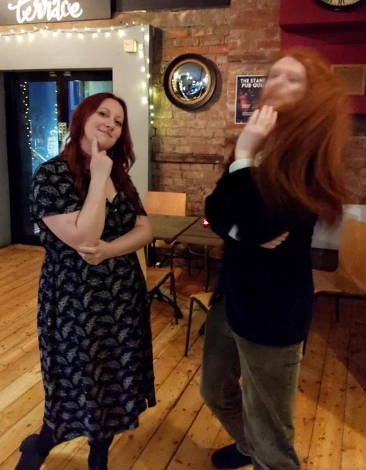 Me on left with long red hair wearing black dress covered in cream moths (a design, not actual moths thankfully). On the right is a man (ABK) with long curly ginger hair, dark blue velvet jacket and khaki corduroy trousers. His face and head is a blur of ginger dynamic movement. Background is the Stand comedy club bar with wooden floor, wooden chairs and brick walls on which is hanging a convex witch's mirror.