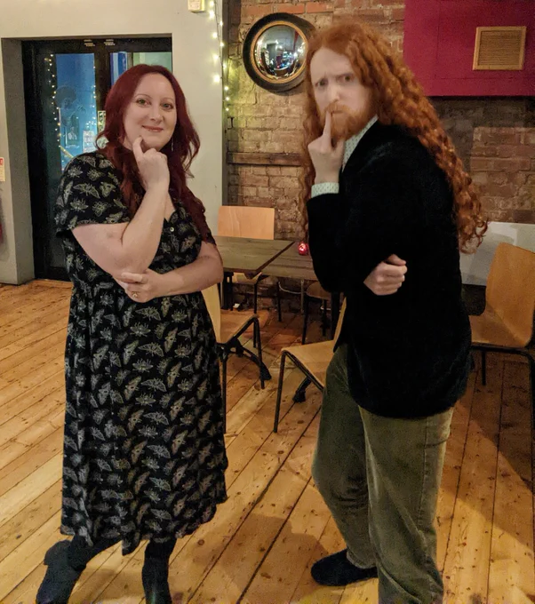 Me on left with long red hair wearing black dress covered in cream moths (a design, not actual moths thankfully). On the right is a man (ABK) with long curly ginger hair, dark blue velvet jacket and khaki corduroy trousers. We have posed our hands in an intellectual way. Background is the Stand comedy club bar with wooden floor, wooden chairs and brick walls on which is hanging a convex witch's mirror.
