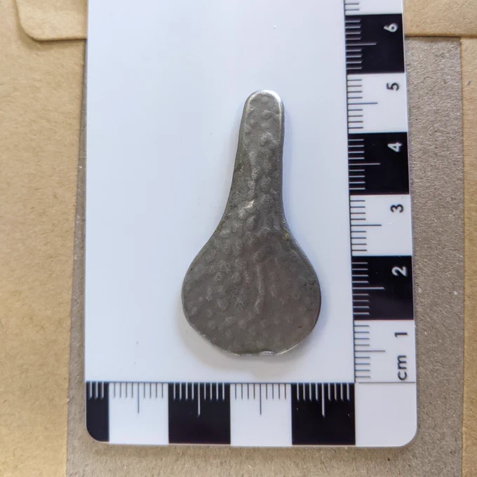 Bike seat shaped white metal object on scale card (approx. 4cm long), dappled finish.