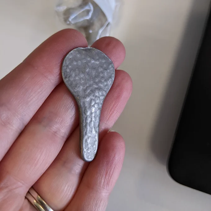 Bike seat shaped white metal object on scale card (approx. 4cm long), dappled finish