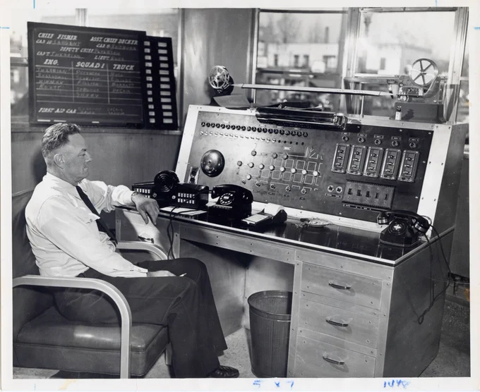 Black and white photo of a man sitting at a desk with multiple telephones and telephone operating equipment on it.