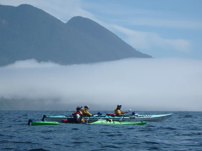 Three young people paddle two kayaks across a calm ocean, with misty mountains in the background. 