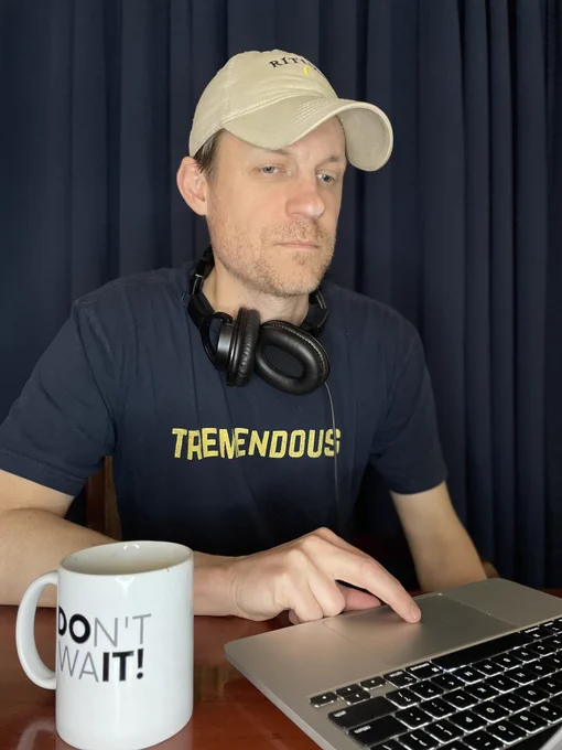 :: IMAGE DESCRIPTION ::
John sitting at a table wearing a navy blue shirt with the word tremendous written in yellow, with his right hand on a laptop trackpad and looking at the screen with his coffee mug nearby with the phrase Don't Wait visible.