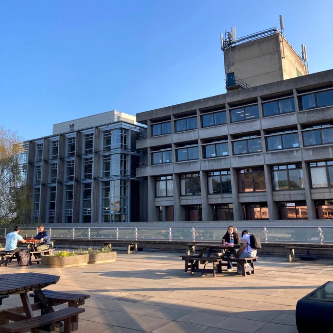 A view of the exterior of the Library in the distance. Ahead of it is an outdoor seating area in the sunshine.