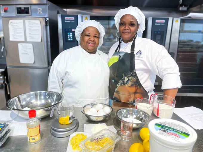 Tekisha Blue and Keisha Acres stand in an industrial kitchen with baking ingredients measured out in front of them. Both are wearing white chef coats and hats.