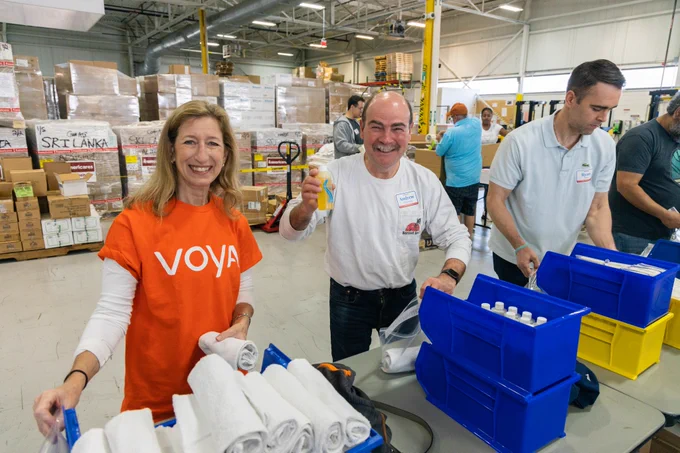A woman in an orange Voya shirt and a man in a white shirt smile while holding items for a hygiene kit, standing at a table behind blue and yellow bins.