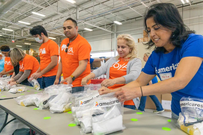 One woman in a blue Americares shirt and five people in orange Voya shirts pack hygiene kits, standing at a table.