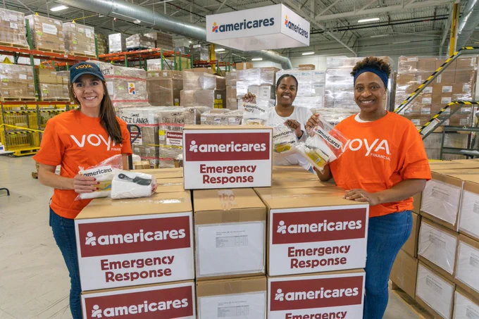 Three women smiling while holding up hygiene kits around a pallet of Americares Emergency Response boxes.