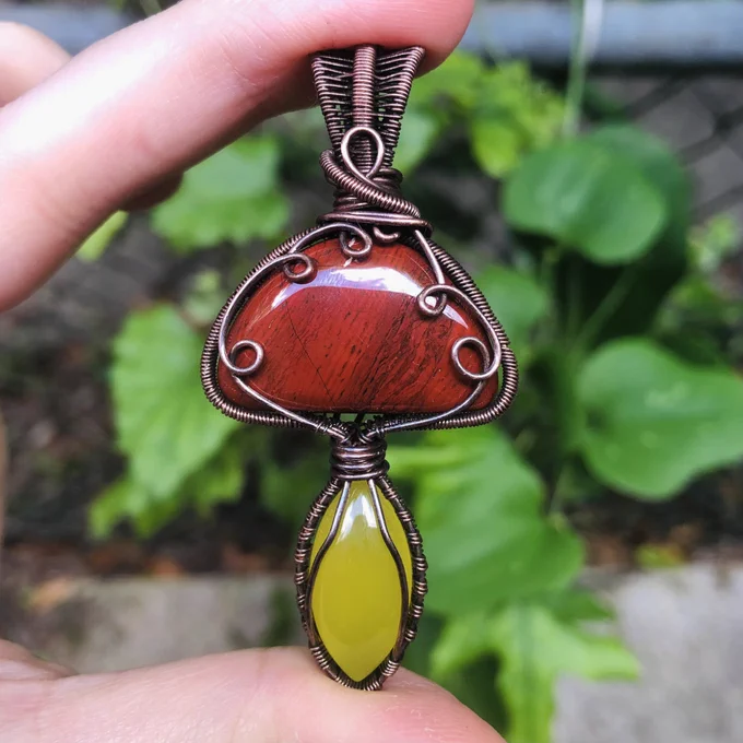 Copper wirewrapped pendant in the shape of a mushroom. The top of the mushroom is red and has swirls and the bottom is yellow. Held in a hand outside in front of green plants.