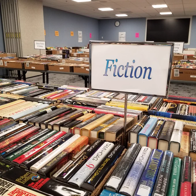 
Used books for sale arranged on tables in a library meeting room. A sign saying "Fiction" is featured.
