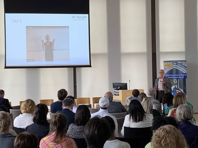 Speaker in front of an audience of people with a presentation in the top left corner.