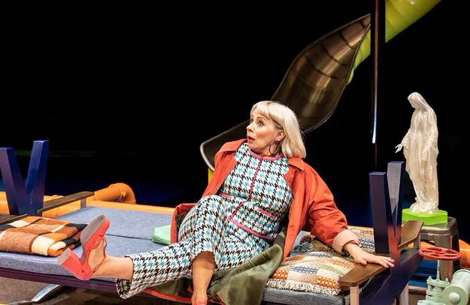 A performer leaning back on a bed onstage, kicking her foot out in front of her. 