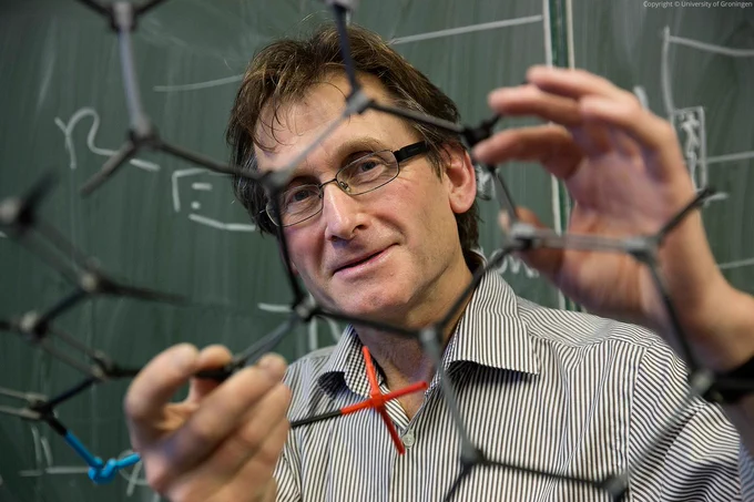 Ben Feringa holding up a molecular model and looking at the camera. He is standing in front of a green board. 