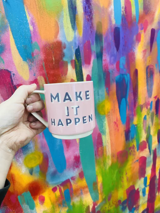 Hand holding a mug with the words ‘Make It Happen’ in front of a colourful wall.