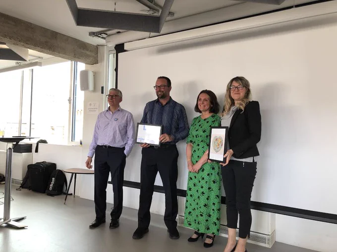 Photo of group of 4 people stood smiling, holding framed paper as acceptance of an award