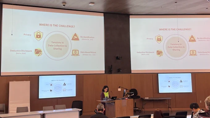 Rie, the speaker, stands at the podium in the Brendan Tribe Center's auditorium. Two large displays are behind her, showing the same slide that outlines the challenge in collecting and sharing data by and about blind users. https://go.umd.edu/datasharing_questionnaire