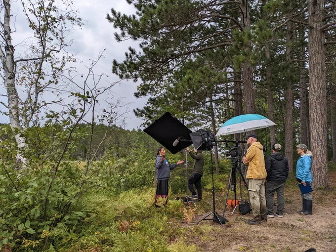 The Clouds North Films film crew interviewing Carolyn L.C. Gouge-Powless of the Red Cliff Band of Lake Superior Ojibwe Kwe. Everyone is standing besides a stand of pines with a wetland area behind them.