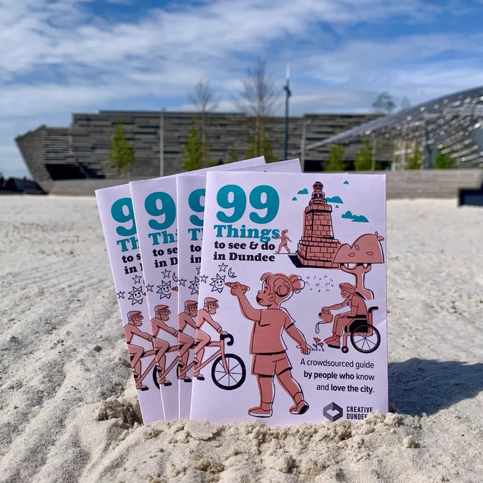 A fan of 4 copies of the 99 Things to See & Do in Dundee guide are propped up in the sand at Dundee's urban beach. The V&A Dundee can be seen in the background.