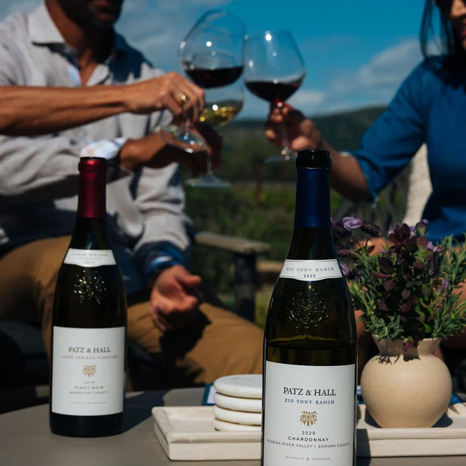 Two bottles of Patz & Hall wine on an outdoor table. One is Pinot Noir and the other is Chardonnay. Three people in the background are holding wine glasses and toasting. 