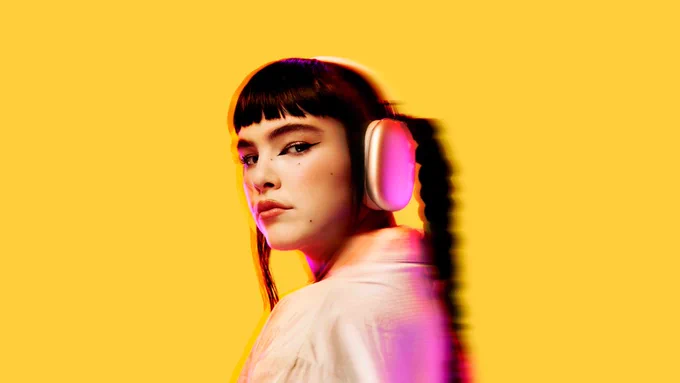 A closeup, pink-lit image of BENEE, wearing a white collared shirt and gold, over-ear headphones, in front of a yellow background.