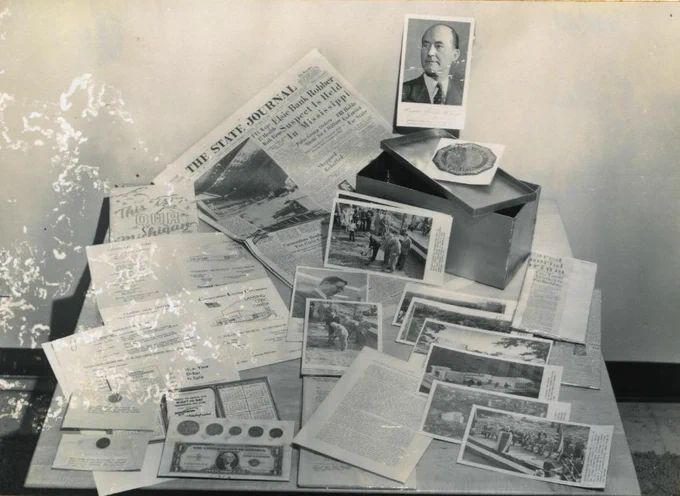 Black and white photo showing the spread-out contents of a time capsule, which includes documents, photographs, a newspaper, and money.