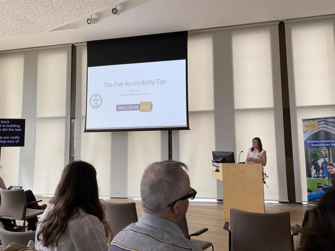 Speaker speaking behind a podium in front of an audience. In the top left centre there is a presentation that says “Top Five Accessibility Tips.”