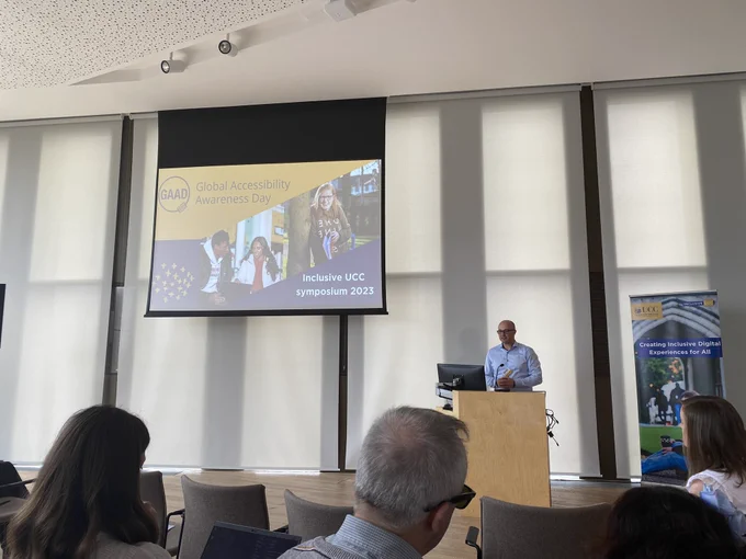 Speaker behind podium speaking in front of a crowd with a projected slide on the top left that says “Global Accessibility Awareness Day - InclusiveUCC symposium 2023.”