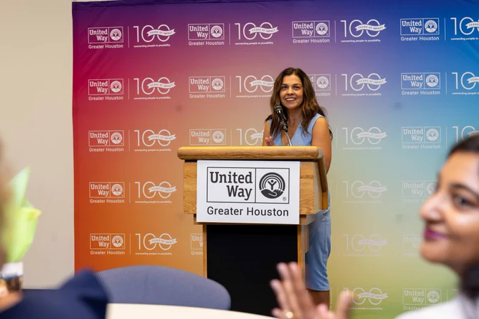 Kiran Handa Gaudioso, wearing a periwinkle sleeveless dress, stands behind a podium with the United Way of Greater Houston logo in front of it and smiles at the crowd in front of her.