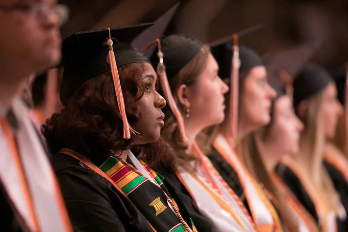 Graduates sit at commencement waiting for the ceremony to proceed. The camera is focused on the upward gaze of a Black woman whose graduate robes include a green, yellow, and red striped stole honoring her participation in the National Society of Black Engineers.