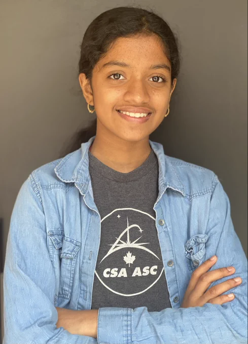 Portrait of Bhavishyaa. Her arms are crossed. She is wearing a grey t-shirt with a Canadian Space Agency logo under a jean blouse. 