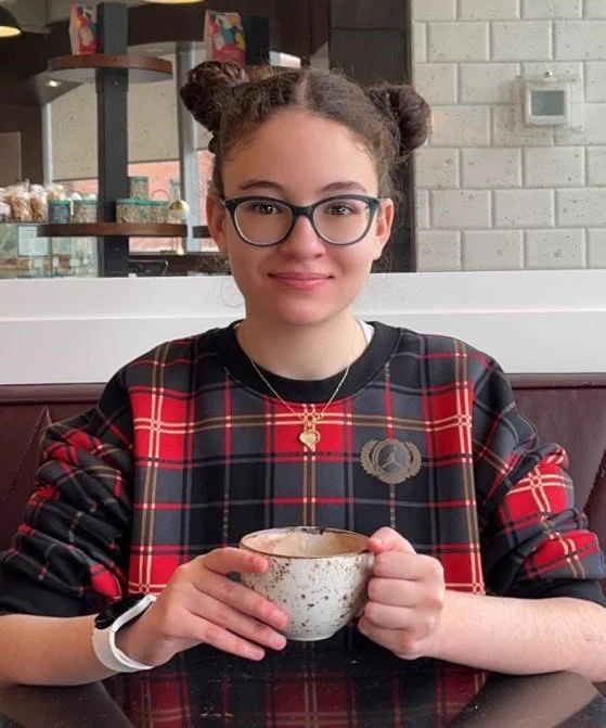 Portrait of Maya. She is sitting at a table holding a mug. 