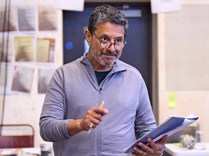 A man in wearing glasses and grey zip up jumper holds a booklet in his left hand and a pencil in his right hand. 
