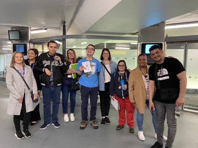 A group of PiP students, staff and HEX colleagues stand in front of the pod parking station. Everyone is standing in a line and smiling.