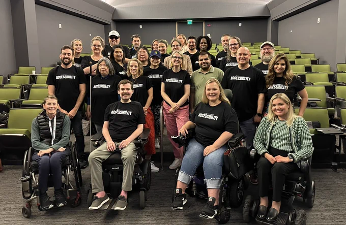 Team of approx 30 people in auditorium, four wheelchairs at the front, many standing with canes, and most wearing black shirt with Microsoft accessibility on the front. Green chairs either side.