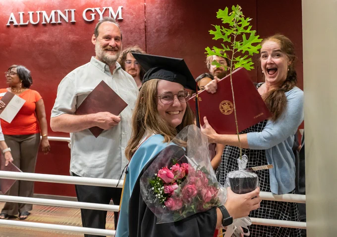 A graduate smiles with her sapling in hand and her family behind her.