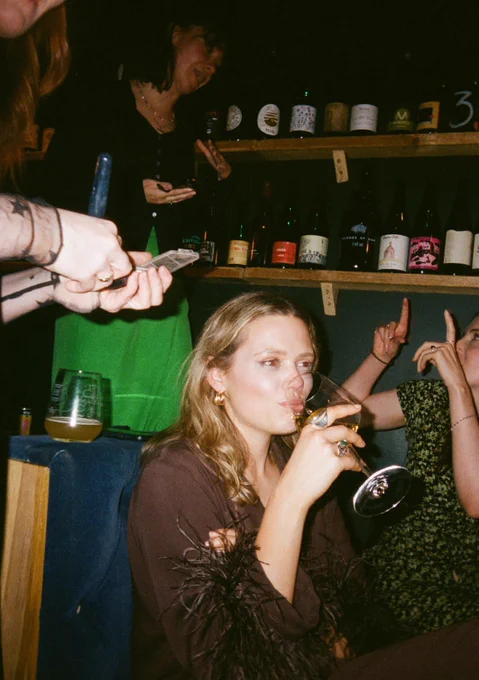 A woman is drinking a glass of wine and looking a bit teary, and people are standing around her with make up.