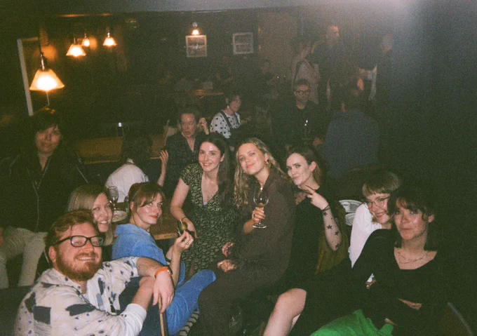 A group of friends are sat in a basement bar, smiling at a camera and holding glasses of wine. Some are sat on each others laps.