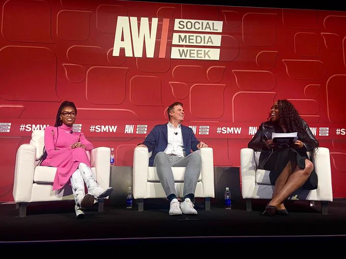 3 people sitting in chairs on a stage in front of a red “Adweek - Social Media Week” background. 
