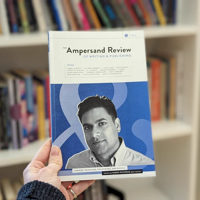 [Image Description: Issue No. 3 of The Ampersand Review is held up in front of a white bookshelf. The bookshelf is filled with many colourful books, but the background is blurred so that no titles are discernable. The cover of Issue No. 3 is in shades of cyan blue, with a grayscale headshot of Naben Ruthnum in the center.]