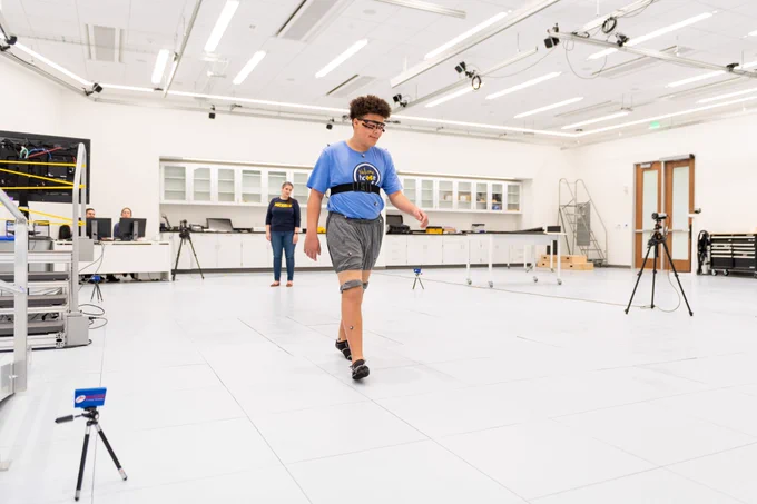 A mixed-race teenage boy wearing athletic clothes, eye-tracking glasses, and motion-capture markers walks across a lab while a white Hispanic woman observes his gait.