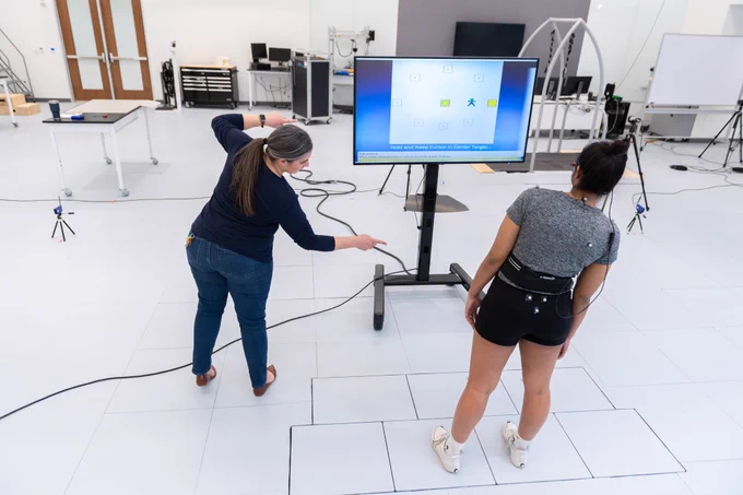 A white Hispanic-American woman demonstrates a leaning task to an Indian-American woman wearing athletic clothing and motion-capture markers while standing in front of a screen displaying a balance game.