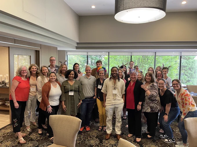 Group photo with over 20 smiling members of NTACT:C Staff. Staff are standing in front of a wall of windows in a hotel restaurant. 