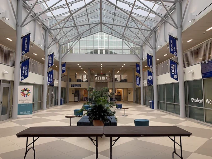 A large atrium of a recently-constructed academic building shows natural light coming through skylights over two stories of offices, with plenty of floor space to line up tables and chairs. 
